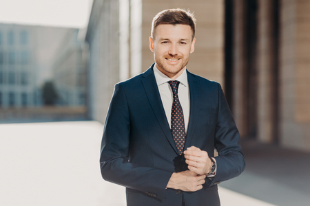 Handsome male with beard, dressed in formal suit, poses outdoor, has watch on arm, being in high spirit after successfully signed contract, feels confident and pleased. Prosperous businessman smilesの写真素材