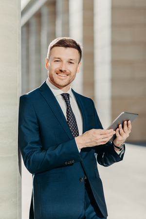 People, work and technology concept. Handsome male economist searches information online on digital tablet, prepares for meeting with staff, works on financial report, dressed in formal clothingの写真素材