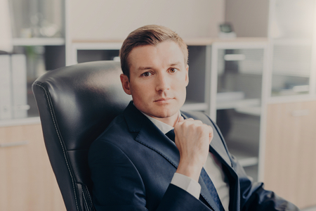Attractive male manager sits in chair at his cabinet, dressed formally, rests for minute after hard work in office. Prosperous man boss has serious expression, works on financial report. Businessの写真素材
