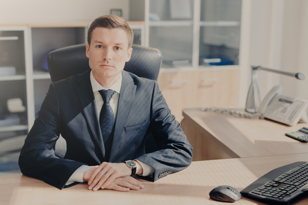Serious male CEO in formal suit sits at work place in cozy cabinet at office, surrounded with necessary technologies and equipment, concentrated on idea for corporation project. Business conceptの写真素材