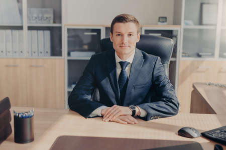 Portrait of attractive prosperous businessman wears black suit, white shirt with tie, sits in his own cabinet, waits for business partners to start meeting, discussing contract terms. Office interiorの写真素材