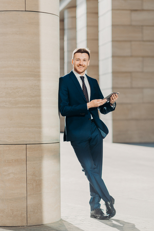 Photo of cheerful male dressed in expensive black suit, stands crossed legs, works on touch pad, stands in modern office interior, smiles positively at camera. Young male manager. Full length portraitの写真素材