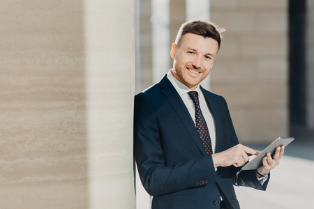 Experienced male economist in formal clothes works on digital tablet, has work break, stands in office interior with copy space. Successful entrepreneur prepares business report on modern deviceの写真素材