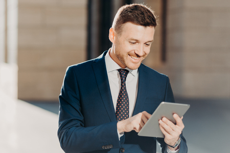 Confident handsome male manager checks email on tablet computer, updates database on touch pad, wears elegant formal clothing, connected to wireless internet, poses outdoor. Business conceptの写真素材