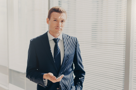 Prosperous male director sends message and multimedia files via smart phone, synchronizes phone data and shares photos for project, poses in office, wears luxury expensive suit, looks seriouslyの写真素材