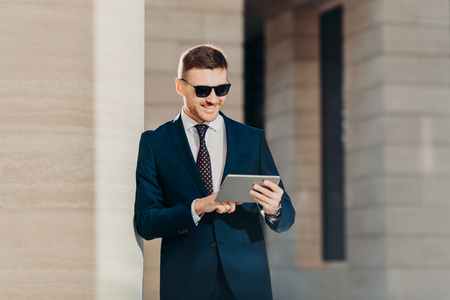 Fashionable young male banker examines future banking agreement, uses modern touch pad, reads important information in internet wears trendy sunglasses and luxury formal suit, smiles gladfullyの写真素材