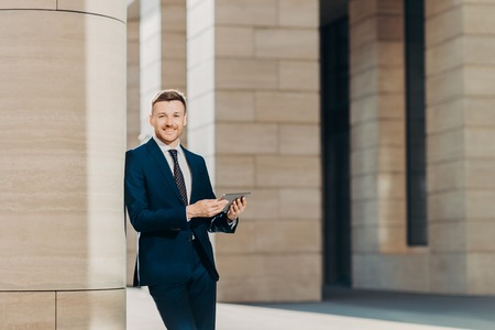 Positive successful male entrepreneur in luxury black suit, holds modern tablet, looks happily somewhere, stands in modern office building, searches internet or social networks during work breakの写真素材