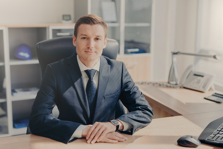 Half length portrait of handsome prosperous male manager sits at cabinet, looks directly into camera, has meeting with colleagues, discuss new business strategy. People and occupation conceptの写真素材