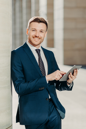Vertical shot of business person uses digital tablet for online communication and work, uses free internet at office, has positive expression, dressed in formal black suit. People and occupationの写真素材