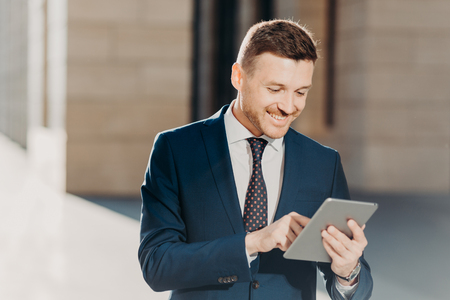 Happy prosperous businessman in formal wear, makes money transaction via touch pad, has glad expression, stands outdoor, connected to wireless intrnet. People, business and technology conceptの写真素材