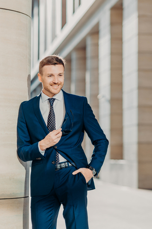 Vertical shot of handsome male with appealing appearance, dressed in formal black suit, keeps hand in pocket, waits for business partners, going to have negotiations, has positive expression.の写真素材