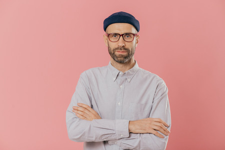 Self confident unshaven man crosses hands over chest, wears spectacles and white shirt, isolated over pink background. Bearded hipster ready for business meeting. People and fashion concept.の写真素材
