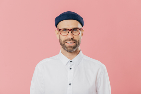 Headshot of satisfied bearded young man looks confidently at camera through spectacles, wears white formal shirt, blue hat, models against pink background, prepares for meeting with investorsの写真素材