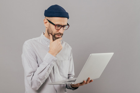 Attentive student holds chin, focused in monitor of laptop computer, searches information for project, connected to wireless internet, isolated over grey studio wall. Networking and people conceptの写真素材