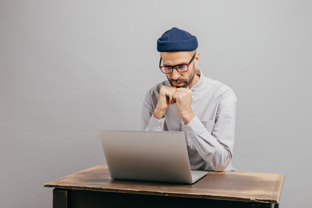 Serious male workaholic sits at desktop with modern electronic device, watches video, prepares financial report, concentrated in laptop computer, wears white shirt, optical spectacles, models indoorの写真素材