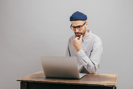 Image of unshaven male owner of trading company searches information about marketing agency, thinks about formal meeting, wears glasses and elegant clothes, sits at old desk over grey backgroundの写真素材