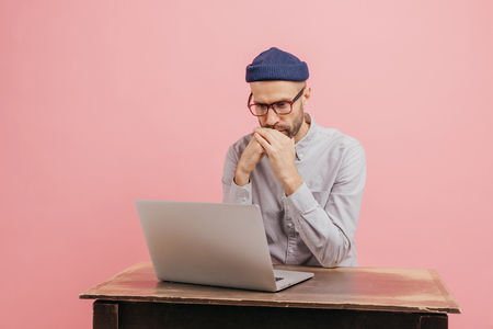 Horizontal shot of concentrated male worker reads statistics attentively, focused in laptop computer, learns something online, wears spectacles, hat and white shirt, isolated over pink wall.の写真素材