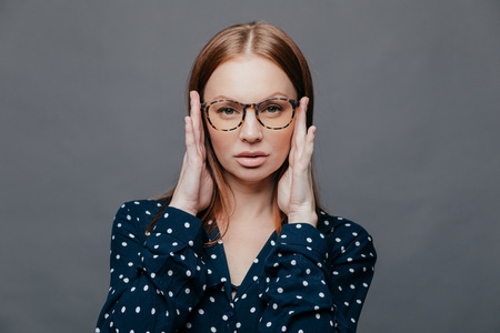 Headshot of serious female teacher keeps hands on head, has self confident facial expression, wears spectacles, polka dot white and black shirt, isolated over grey background. Horizontal shotの写真素材