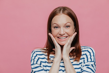 Headshot of attractive young woman touches cheeks with both palms, has sincere smile, wears casual white and blue striped sweater, isolated over pink background with free space for your textの写真素材