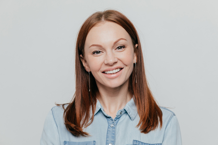 Photo of attractive young woman with dark straight hair, smiles happily, dressed in denim shirt, poses over white background, expresses positive emotions, models indoor. People, beauty, lifestyleの写真素材