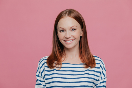 Headshot of pretty smiling European woman with charming smile, wears striped jumper, has brown hair, looks directly at camera, isolated over pink studio background. People, joy, happiness conceptの写真素材