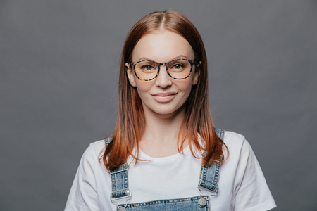 Happy young woman with glad facial expression, has healthy skin, wears optical glasses, white t shirt and denim sarafan, models over grey background, going to meet with friends, poses indoorの写真素材