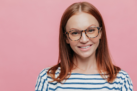 Close up shot of adorable young female student wears optical glasses, has tender smile, rejoices holidays wears striped clothes, models over pink background with copy space for your information.の写真素材