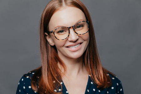 Cheerful close up shot of pretty woman with brown hair, toothy smile, wears spectacles, dressed in formal blouse, poses over grey background, being pleased to meet with colleagues on meetingの写真素材