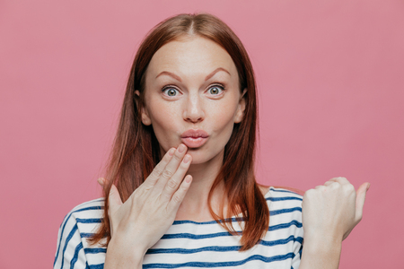 Close up shot of attractive woman raises eyebrows, pouts lips, keeps hand near mouth, has brown hair, surprised by something, models over pink background. People, facial expressions and beauty conceptの写真素材