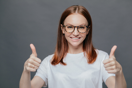 Glad positive woman with tender smile on face, has brown hair, raises two thumbs, demonstrates her approval, isolated over grey background, wears spectacles, wears casual white t shirt. Body languageの写真素材