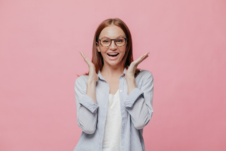 Studio shot of joyful excited woman dressed in striped shirt, spreads hands near face, wears optical glasses, models over pink background, doesnt expect to notice something pleasant and unexpectableの写真素材