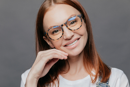 Close up portrait of pretty female model with European appearance, smiles gently, glad to recieve good comments from boss, wears spectacles, keeps hand near face, isolated over grey wall, tilts headの写真素材