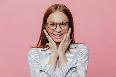 Photo of atractive young woman teacher with toothy smile, touches cheeks, happy to recieve praise, wears formal shirt, likes her profession, expresses happiness, isolated over pink backgroundの写真素材