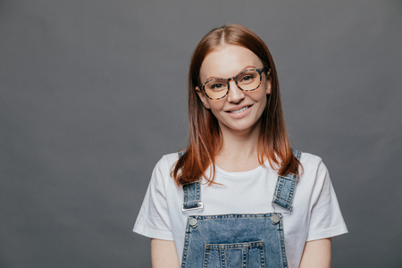Isolated shot of positive pretty woman tilts head, has charming smile, wears glasses, white t shirt and overalls, poses over grey background with free space for your advertising content or promotionの写真素材