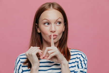 Pretty curious Caucasian woman makes silence sign, keeps index finger over mouth, looks secretly aside, wears striped casual clothes, poses over pink studio wall with free space for your text. Hushの写真素材