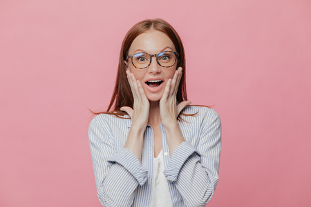 Omg, how nice! Beautiful elegant female employee touches cheeks with both palms, wears spectacles, elegant shirt, cant believe in her promotion, recieves reward, isolated over pink backgroundの写真素材