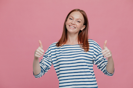 Positive young European woman with reddish hair keeps two thumbs raised, tilts head, dressed in striped clothes, demonstrates agree gesture, wears striped sweater, isolated over pink backgroundの写真素材