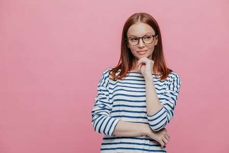 Waist up shot of attractive young woman holds chin, keeps hands partly crossed, being deep in thoughts, stands against pink background with copy space for your promotional content or advertisementの写真素材