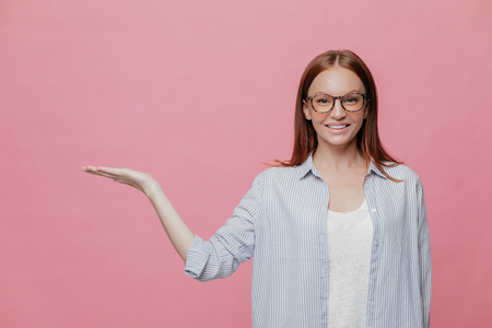 Indoor shot of European woman with pleasant appearance, charming smile, holds copy space, raises palm, wears transparrent glasses, being in high spirit, isolated over pink background. Advertisementの写真素材