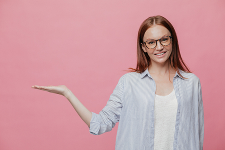 Pleased young woman in formalwear keeps palm raised, being in good mood, advertises something against blank copy space, has satisfied expression, isolated over pink background, gestures indoorの写真素材