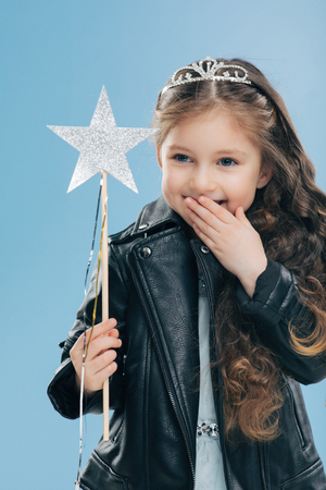 Glad pleasant looking small kid covers mouth with palm, giggles positively, wears crown and black leather jacket, holds magic wand, isolated over blue background. Children and happiness conceptの写真素材