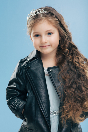 Isolated shot of beautiful Caucasian small child has blue eyes, long dark hair, keeps hands in pockets of leather jacket, isolated over blue background. Children, style, childhood, fashion conceptの写真素材
