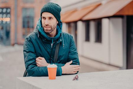 Pensive unshaven young Caucasian man with bristle, wears hat and jacket with hoody, drinks takeaway...