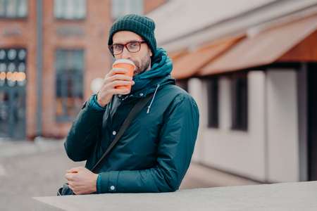 Outdoor shot of handsome hipster drinks takeawy coffee from disposable cup, dressed in headgear, jacket, wears spectacles, has srtoll outside, has straight look at camera. People and lifestyleの写真素材