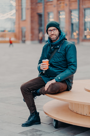 Concept of drinks and lifestyle. Carefree satisfied European man with bristle enjoys sunny day and hot beverage, smiles happily, sits against street background. Outdoor shot of handsome hipsterの写真素材