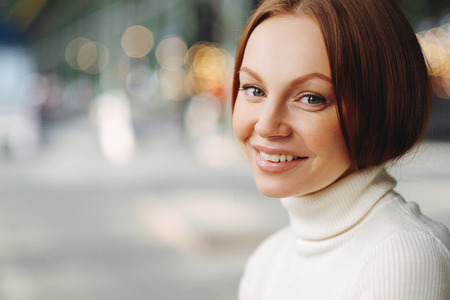 Sideways shot of pleasant looking woman with make up, tender smile, dressed in white turtleneck sweater, stands against blurred background with copy space for your advertisement, has healthy skinの写真素材