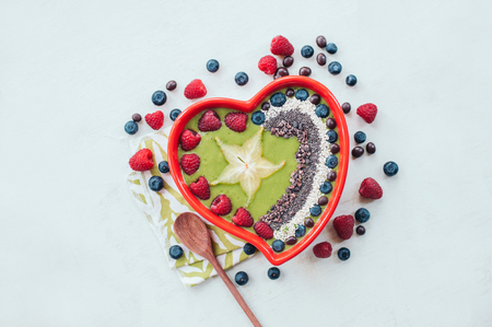 Heart shaped bowl of raspberry, blueberry, cocao nibs, wooden spoon near, isolated over white backgound. Spinach bowl. Fresh fruitの写真素材