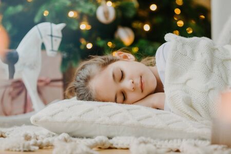 Close up shot of attractive small child lies on floor, sleeps near New Year or Christmas tree, warms with white knitted sweater, has pleasant dreams in evening. Children, winter holidays, rest conceptの写真素材