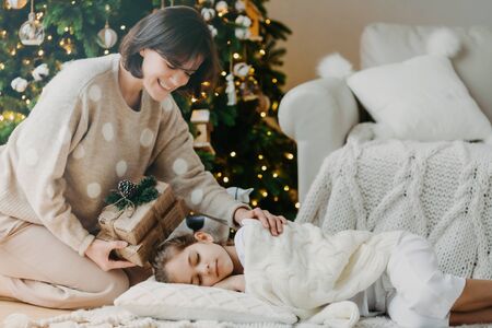 Famaily, happy winter holidays concept. Caring mom stands on knees near sleeping daughter, prepares presents for New Year, has pleasant smile, pose in cozy living room. Christmas night and miracleの写真素材