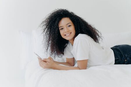 Pretty cheerful woman with Afro haircut, enjoys chatting and networking, lies on comfortable bed, looks photos, has relaxed face expression, isolated over white background. Online communicationの写真素材